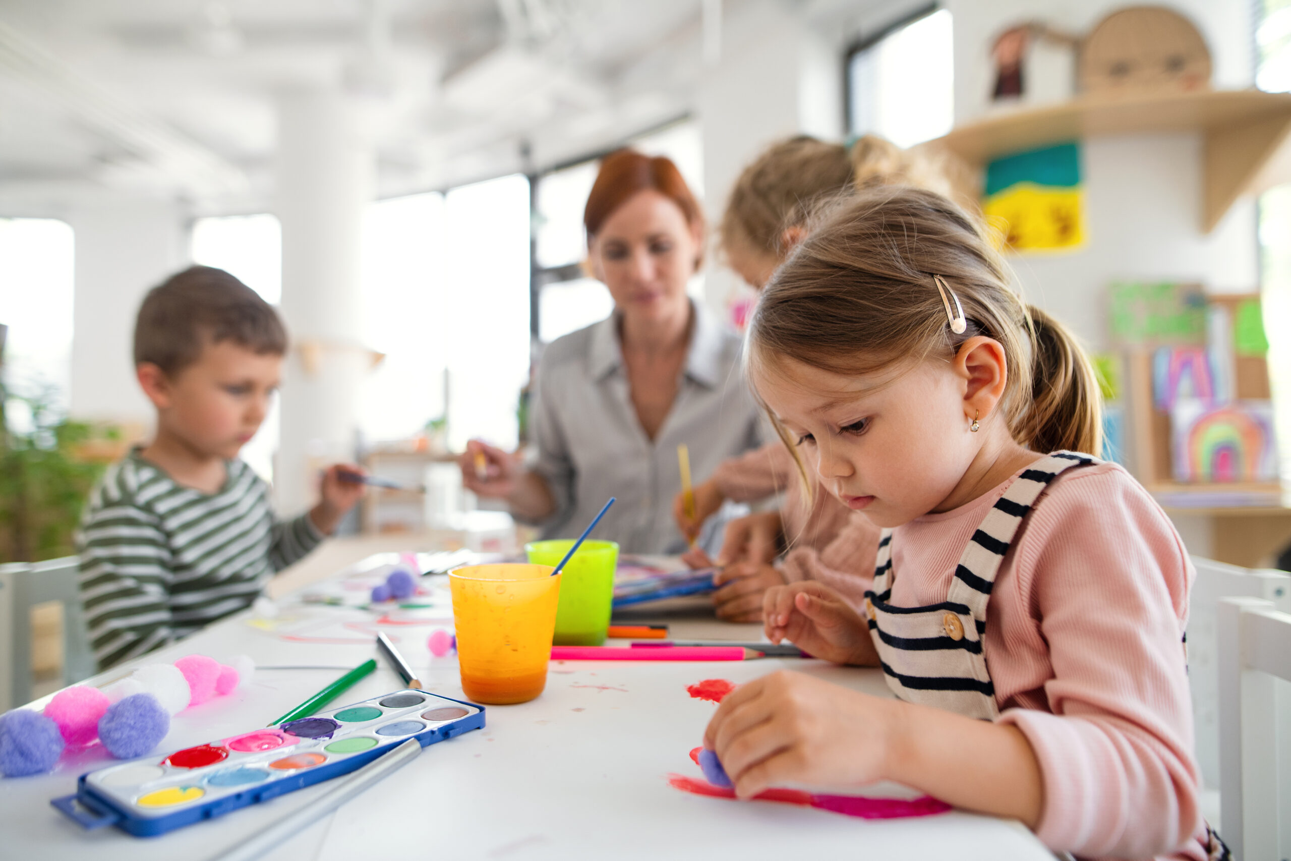 Group of small nursery school children with teacher indoors in classroom, painting. 3 enfants en bas âge, et une assistante maternelle TNS, dans une crèche